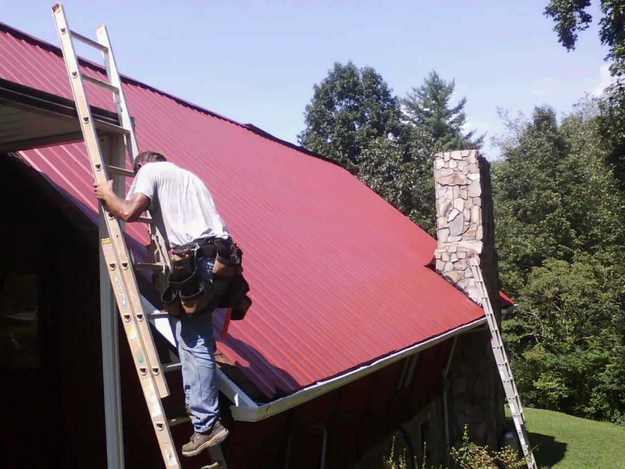 Roofer on a ladder beside a red metal roof and stone chimney — Soffit & Fascia Repair by Roofing Brothers in Chewelah
